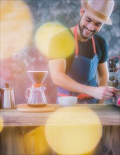 Cheerful man in a blue apron grinds coffee beans, bathed in sunlight with bright yellow orbs,