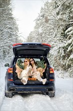 Two women are sitting in the trunk of a car holding in hands bouquets of fir branches and wine