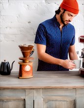 Focused barista in a blue patterned shirt and red beanie uses a manual coffee grinder, Vertical