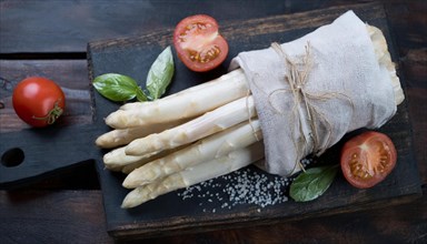 Fresh asparagus bundled on a wooden board next to tomatoes and basil, bunch of white asparagus