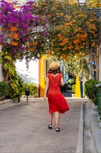 A woman in a red dress walks down a street with a hat on. The street is lined with colorful