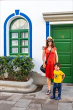 A woman and a child stand in front of a green door. The woman is wearing a red dress and a hat