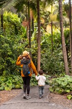 A woman and a child are walking through a forest. The woman is wearing a yellow hat and a backpack.