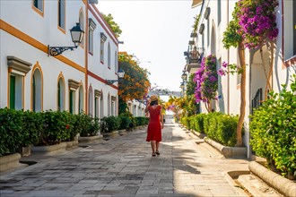 A woman in a red dress walks down a narrow street lined with trees and buildings. The street is