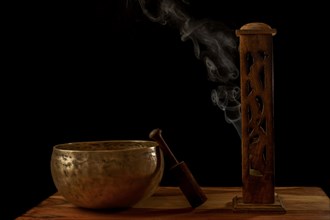 Tibetan singing bowl next to a wooden censer with smoke coming out isolated on black background