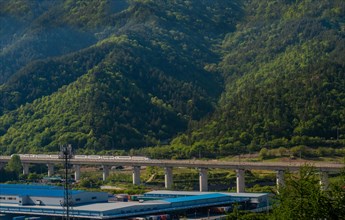Train bridge spanning over an area with industrial facilities against a backdrop of mountains, in