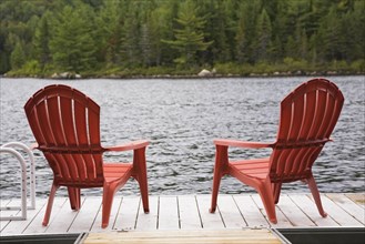 Two bright red plastic Adirondack chairs on wooden floating dock on calm lake with forest of green