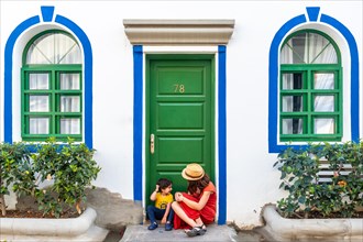 A woman and a child are sitting on the steps of a building with a green door. The woman is wearing
