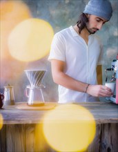 A bearded man in a knit hat prepares a coffee with a machine, soft yellow lights blurred in the
