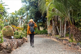 A woman is walking down a path in a garden. She is wearing a yellow jacket and a hat. She is