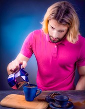 A caucasian man pouring coffee into a blue mug, standing over a wooden board, looks contemplative,