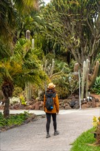 A woman wearing a hat and a backpack walks through a garden. The woman is walking on a path