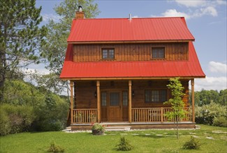 Small two story brown stained log cabin home with veranda and red sheet metal roof and landscaped