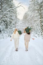 Rear view of two women walking in snowy forest holding small Christmas trees on their shoulders