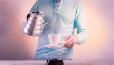 A man in a pastel sweater manually brewing coffee by pouring from a kettle into a ceramic cup,