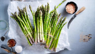 Fresh green asparagus lies bundled next to sea salt and cooking utensils on a wooden board, KI
