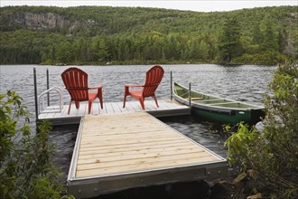Two bright red plastic Adirondack chairs on floating wooden dock and green rowboat on calm lake