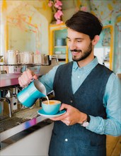 A smiling bearded barista pouring milk into a coffee cup, standing by an espresso machine inside a