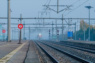 Desolate railway station in foggy conditions at dawn with signal lights on and a train approaching,