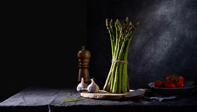 A bunch of green asparagus stands next to a pepper mill and fresh vegetables, fresh green