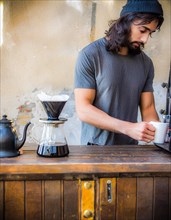An earnest barista in a black t-shirt brews coffee on a wooden counter with a modern, minimalistic