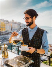 Man brews coffee outdoors using a pourover method with a breathtaking sea view and clear blue sky,