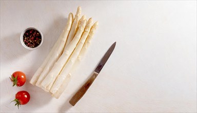 Fresh white asparagus next to tomatoes, a pepper mill and a knife on a light-coloured surface, AI