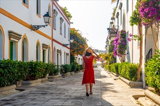 A woman in a red dress walks down a narrow street with a hat on. The street is lined with trees and