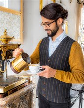 An elegant man in a vest pours coffee from a golden pot in a luxurious, traditionally adorned room,