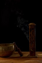 Tibetan singing bowl next to a wooden censer with smoke coming out isolated on black background