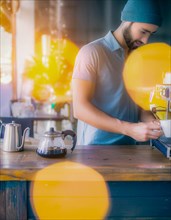 Man focuses while preparing a pour-over coffee with kettle on a wooden surface, yellow bokeh lights