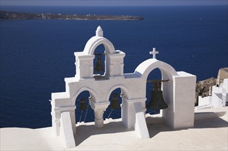 White roughcast cladded arched church bells tower with cross overlooking the Aegean sea, Oia