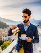 A casually elegant wealthy dressed man pours coffee on a balcony overlooking the city in the