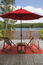 Red canvas cloth parasol and wooden folding canvas chairs on red mat on grey wood plank deck with