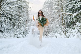 Playful woman kicking snow in winter forest holding glass of champagne and fir branches in hand
