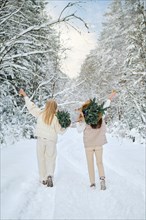 Rear view of two happy women walking in winter forest carrying small Christmas trees on their