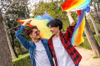 Multiracial gay couple raising lgbt rainbow hand fan smiling walking in a park