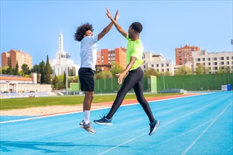 Happy friends african american young runners jumping high-fiving in an outdoor blue running track