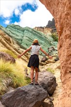 A tourist woman with a hat and open arms at the Azulejos de Veneguera or Rainbow Rocks in Mogan,