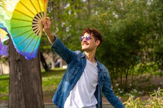 Happy caucasian casual gay man dancing with rainbow fan in a park