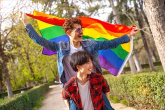 Multiracial gay couple waving lgbt flag piggybacking celebrating diversity in an urban park