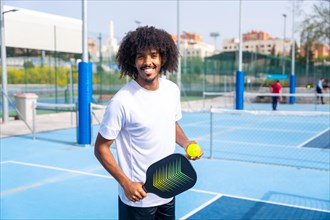Portrait with a man with afro hairstyle smiling in a pickleball court holding a racket and ball