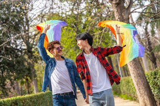 Happy multiracial gay couple walking holding hands and waving LGBT rainbow hand fans in a park
