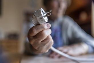 Senior citizen holding a power cable with plug in her hand at home, symbolising energy costs and