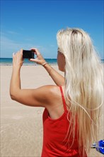 Blonde woman in a red dress taking a photo with her smartphone on a sunny beach