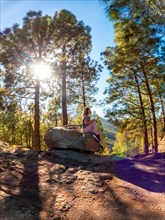A woman sits on a rock in a forest. The sun is shining on her, creating a warm and peaceful
