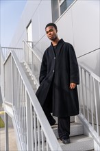 Vertical portrait of an African beauty man posing with long black jacket in stairs