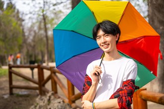 Asian gay man posing with rainbow umbrella standing in a park smiling at camera
