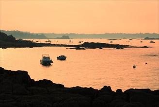 Calm sea with boats and rocks silhouetted against an orange sunset sky