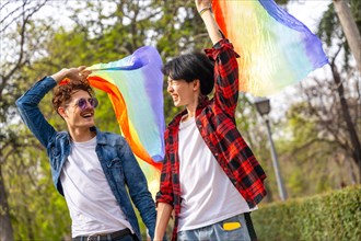 Happy multi-ethnic gay men holding hands raising lgbt flags walking along a park